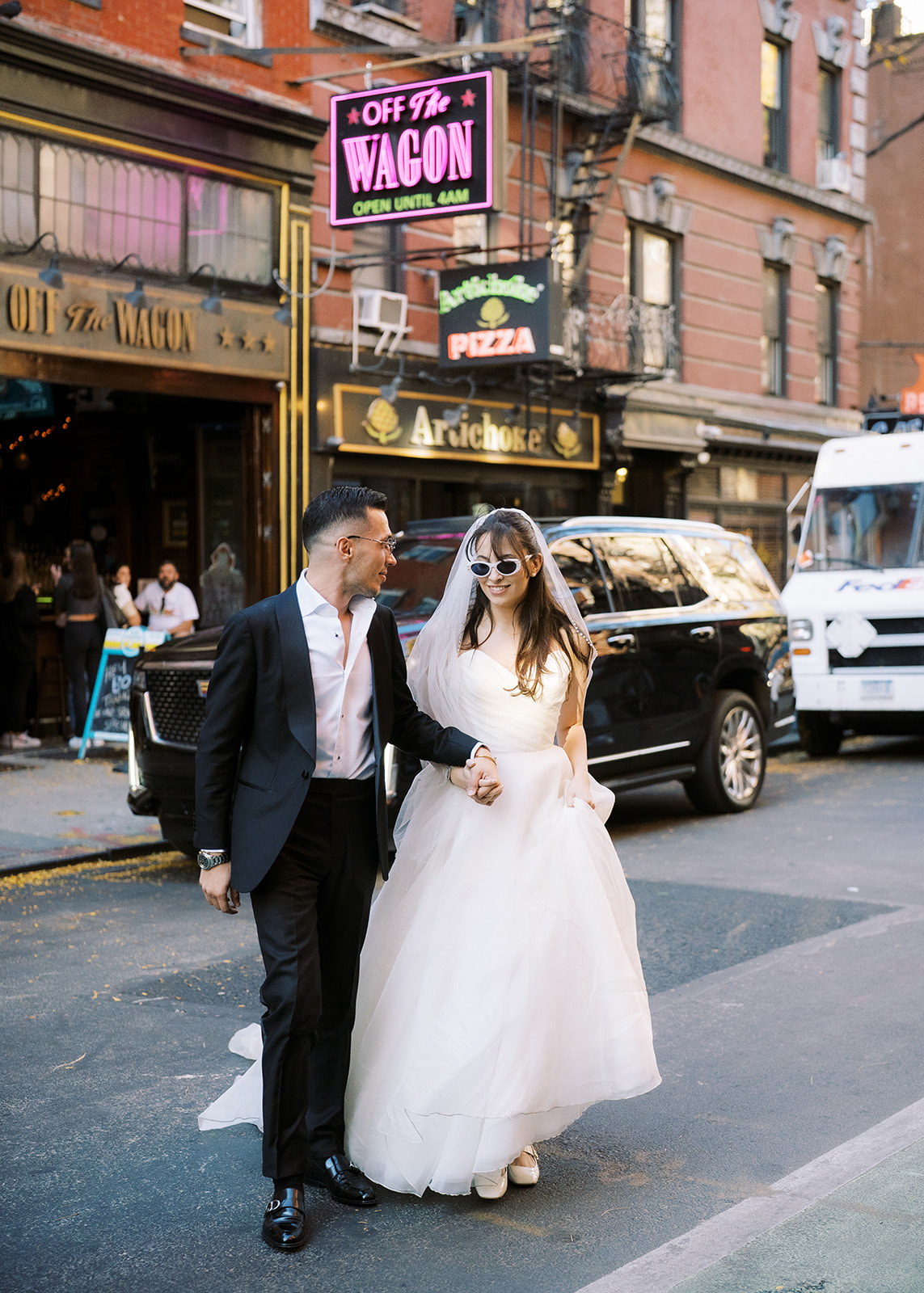 bride and groom crossing the street in the west village with artichoke pizza in the background and wearing sunglasses